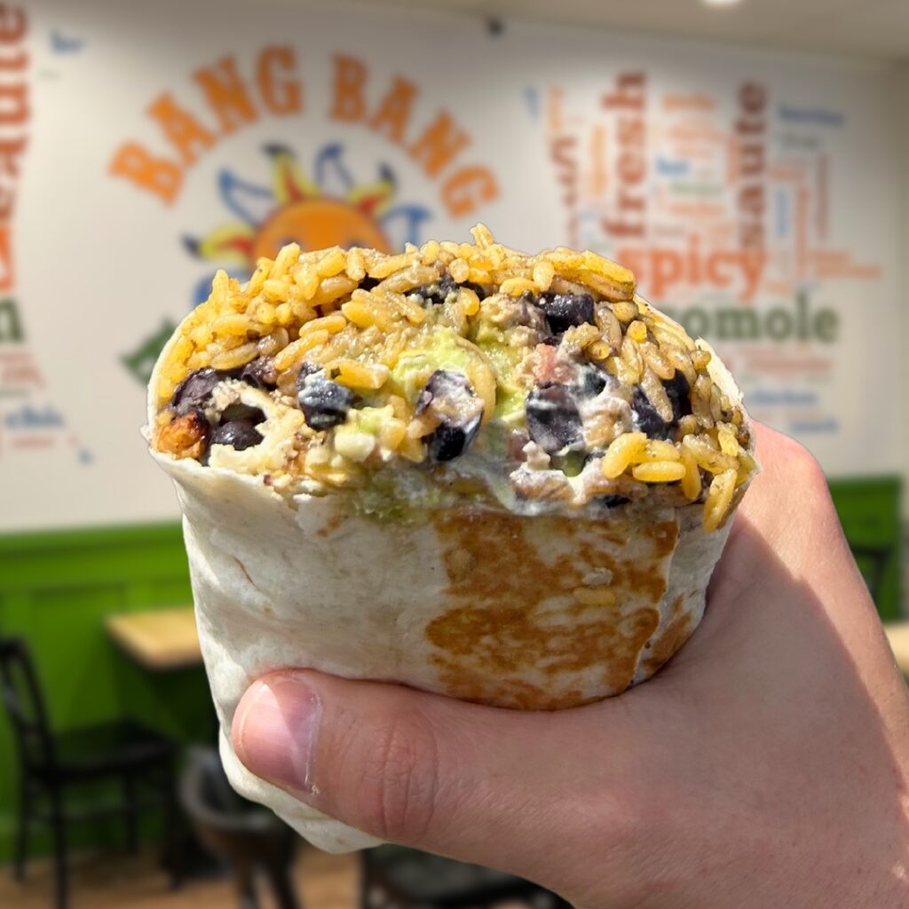 A hand holding a halved Bang Bang Burritos burrito filled with rice and black beans inside the restaurant